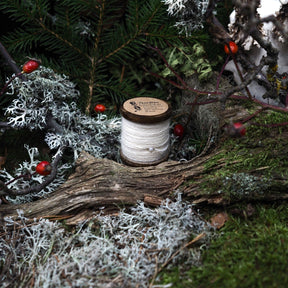 Naturvit brodyrtråd i 100% ull på träspole, placerad på naturlig mosslik miljö. Cirka 50 meter tråd för broderi och traditionellt handarbete. Fotograferad i utomhusmiljö.
