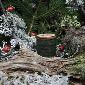 Green embroidery thread on wooden spool placed in a natural forest scene with moss, berries and ancient atmosphere