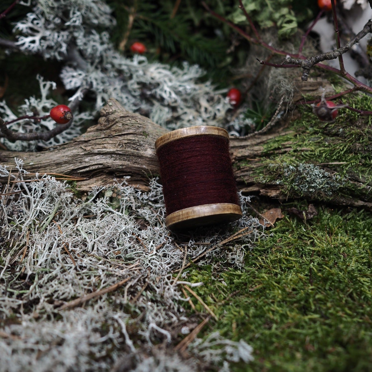 Djupt röd brodyrtråd 100% ull på traspole i naturlig miljö, visar produktens traditionella handverksaspekt