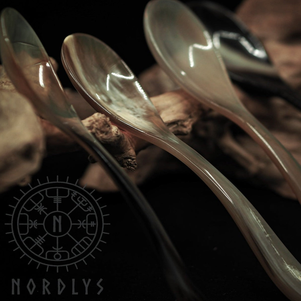 Matsked i horn, flerfärgad variant. Närbild visar flera horn-matskedar med naturlig amber-guldfärgning, genomskinliga striningar och graverad Nordlys-logo med vinkingoriginspiration, fotad mot mörk bakgrund.