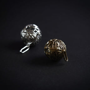 Två berlocker med livets träd-mönster i silverpläterad och mässing-finish, ligger bredvid varandra för att visa material-skillnaden. Båda har samma intrika skärning av trädgrenar och rötter.