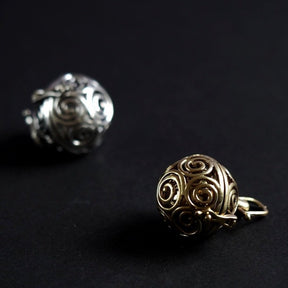 Två berlocker med solspiralmönster - en i silverpläterad finish och en i mässing. Båda visar samma intrika spiraldesign utskärd genom den sfäriska bärlocken, fotograferad från sidan för att visa mönstrets komplexitet.