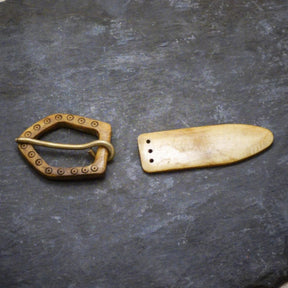 Två delar av ett bältesspänne i ben: en D-formad spänne med dekorativ prickad rand och en långsträckt bältesände med två fästhål, presenterat på mörk bakgrund