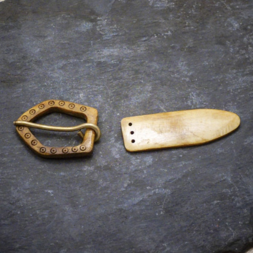 Två delar av ett bältesspänne i ben: en D-formad spänne med dekorativ prickad rand och en långsträckt bältesände med två fästhål, presenterat på mörk bakgrund