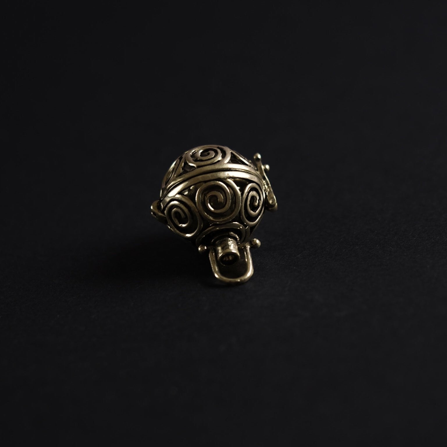 En mässing-berlock med spiralmönster utskärt i sfärisk form, fotograferad närgånget mot mörk bakgrund. Designen visar solspiraler - ett klassiskt vikingamotiv - och bärlocken är gjord i mässing för en varm, gyllene ton.