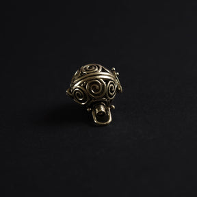 En mässing-berlock med spiralmönster utskärt i sfärisk form, fotograferad närgånget mot mörk bakgrund. Designen visar solspiraler - ett klassiskt vikingamotiv - och bärlocken är gjord i mässing för en varm, gyllene ton.