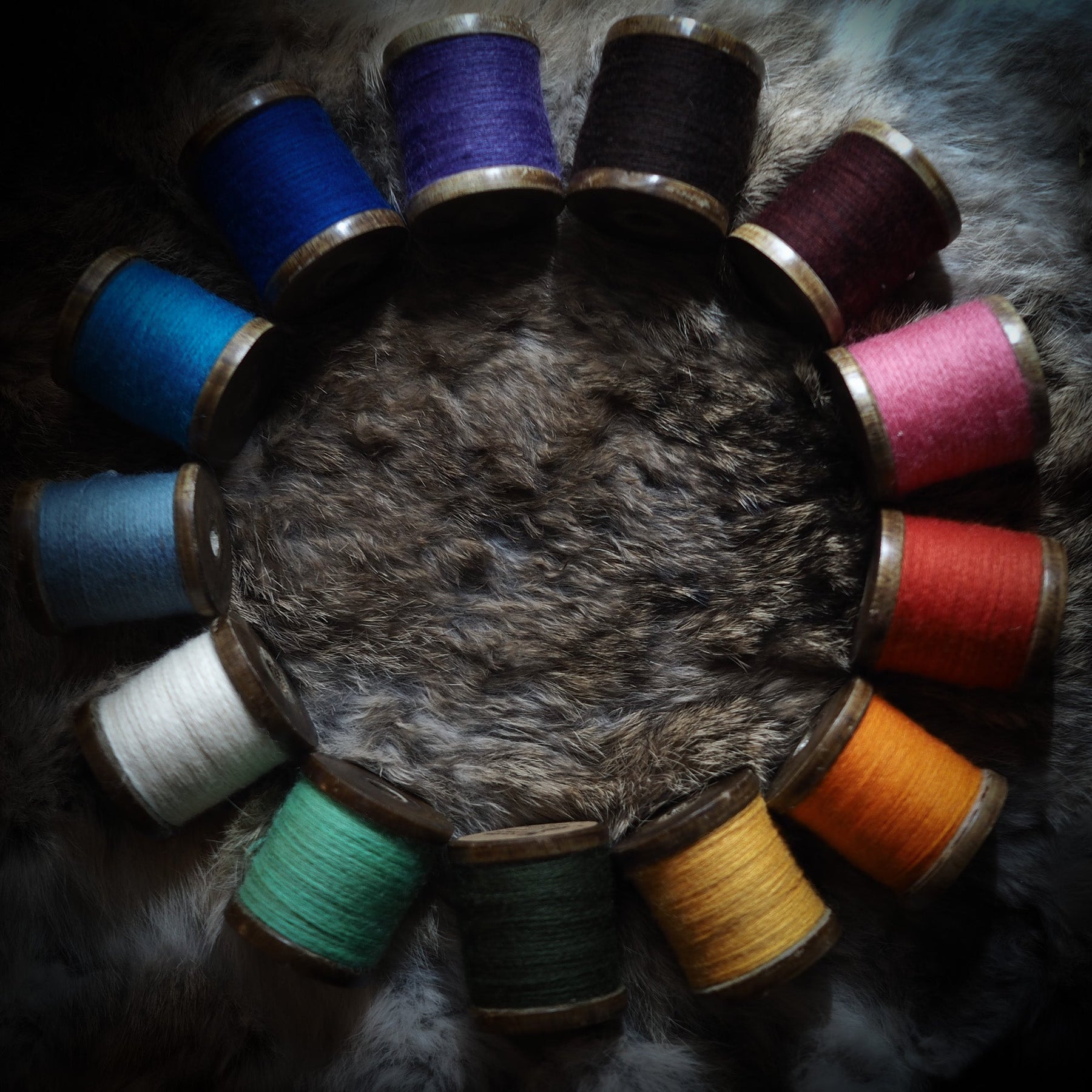 Tolv farvede broderitråd-spoler arrangeret i cirkel omkring dyrehud på mørk baggrund, viser traditionel nordisk tekstilkunst