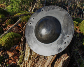 Metallbuckler sköld från ovan på mossbevuxen skogsmark, visar konvex bål och silvergrå ram