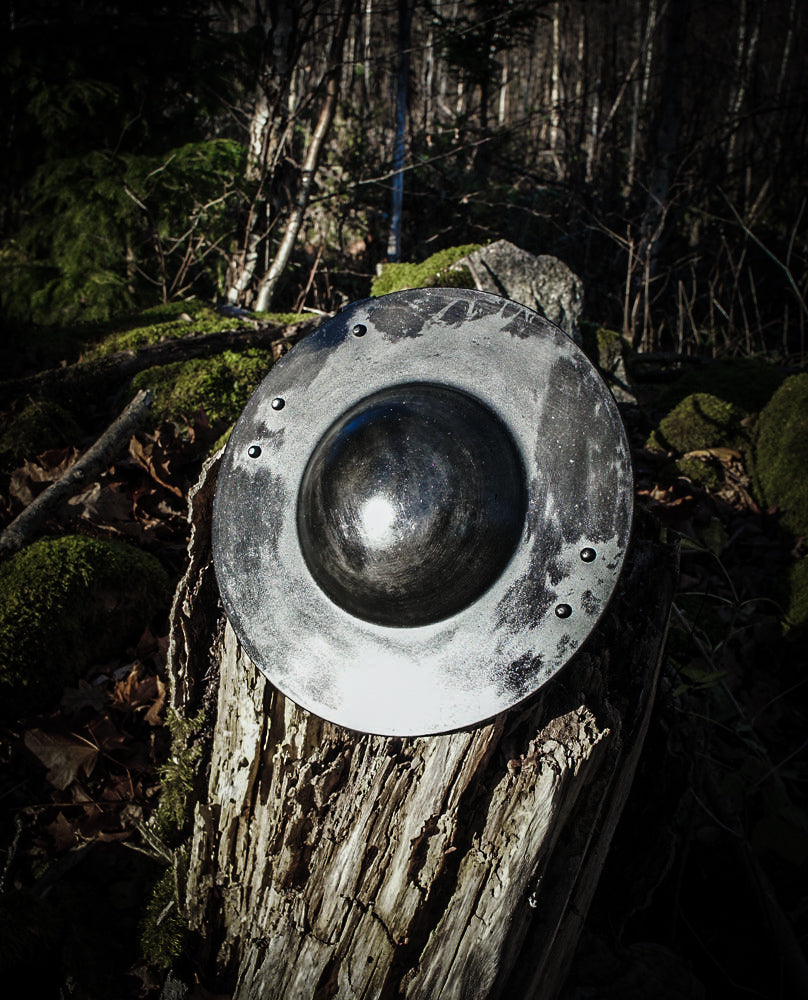 Metallbuckler framifrån fotograferad från ovan på skogsmark med moss och löv, visar konvex bål och rivetnitar
