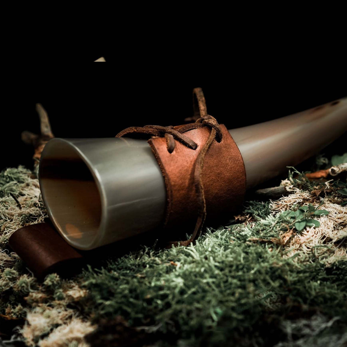 Hornhållare av brunt läder med organiskt horn infört i cylindrisk läderväska, på mörk bakgrund med grön mossa