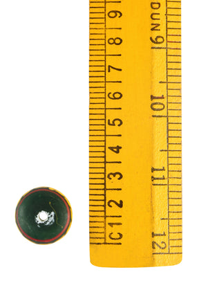 Stor grön glaspärla fotograferad mot gul måttstav för referens. Pärlans diameter cirka 15-16 mm. Röda och gula dekorativa mönster i vågig stil. Visar vita slitmarkeringar. Dokumentationsfoto av arkeologisk typ från Bodarp, Skåne, Sverige.