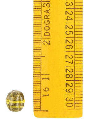En melonpärla av glas med gul mässingsomslutning framför en gul måttstock för skalreferens, visar pärlans små dimensioner omkring 9-10mm