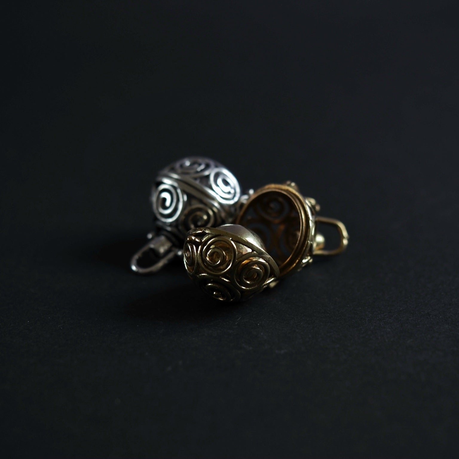 Tre berlocker med solspiralmönster visade i olika materialfinisher - silverpläterad och två mässing-varianter. Foto visar materialskillnaderna mellan silverpläterad (sval, silvrig glans) och mässing (varm, gyllene ton). Alla har identisk spiral-skärningsdesign.