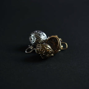 Tre berlocker med solspiralmönster visade i olika materialfinisher - silverpläterad och två mässing-varianter. Foto visar materialskillnaderna mellan silverpläterad (sval, silvrig glans) och mässing (varm, gyllene ton). Alla har identisk spiral-skärningsdesign.