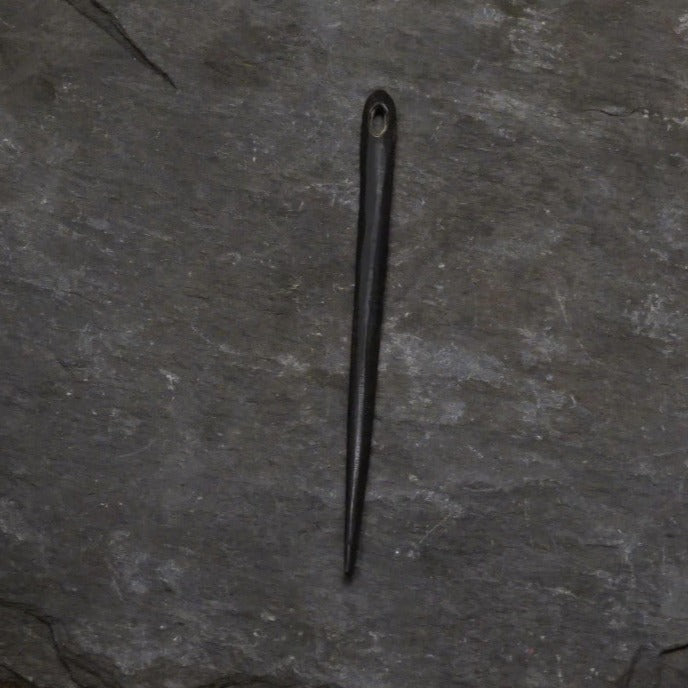 Nålbindingsnål av horn i svarttonart, fotograferad på naturlig skogsmarksbakgrund med mossa och växtliv