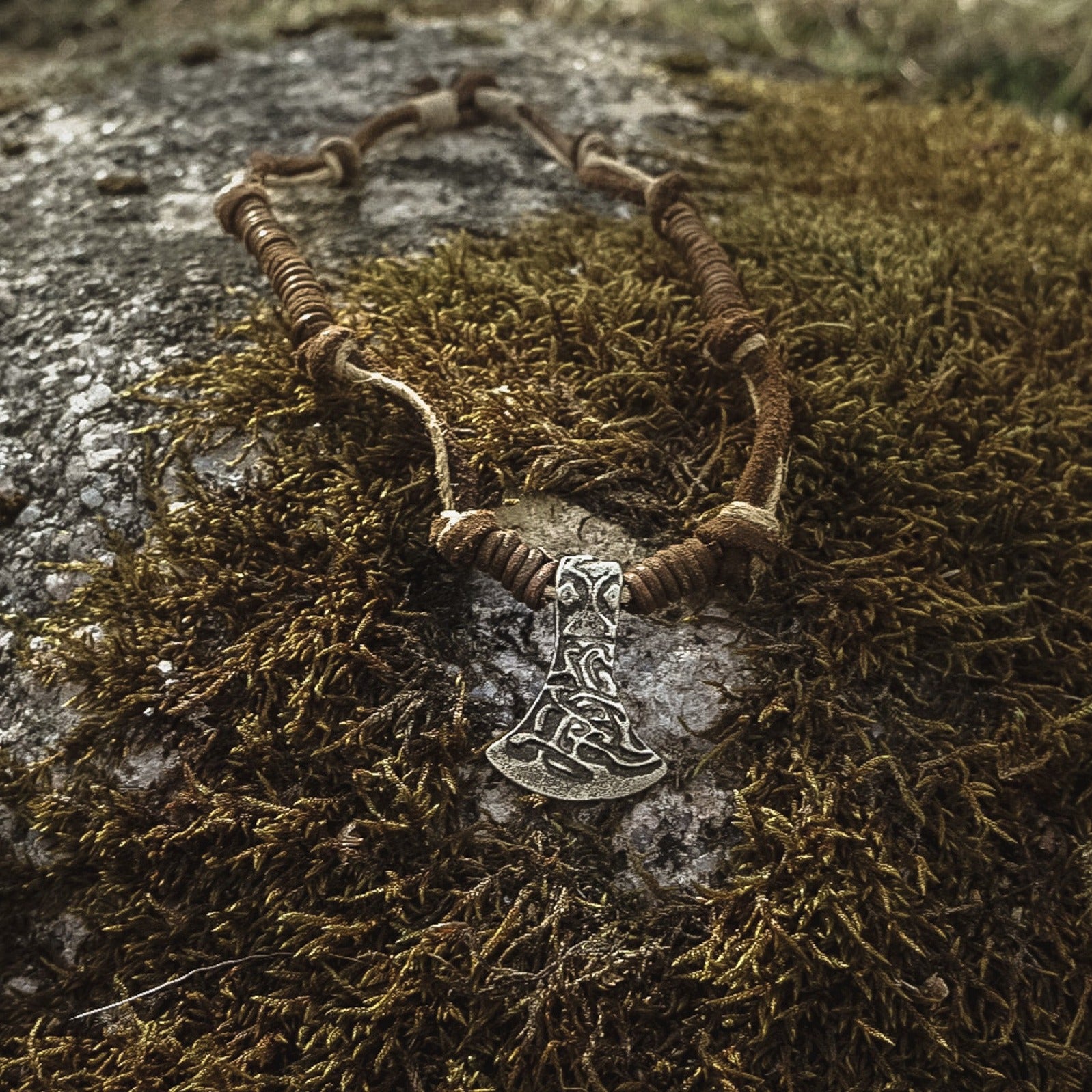 En halsband med läderband och en tenn-ax på en mossig sten. Naturlig omgivning med axans ornerade mönster synligt.