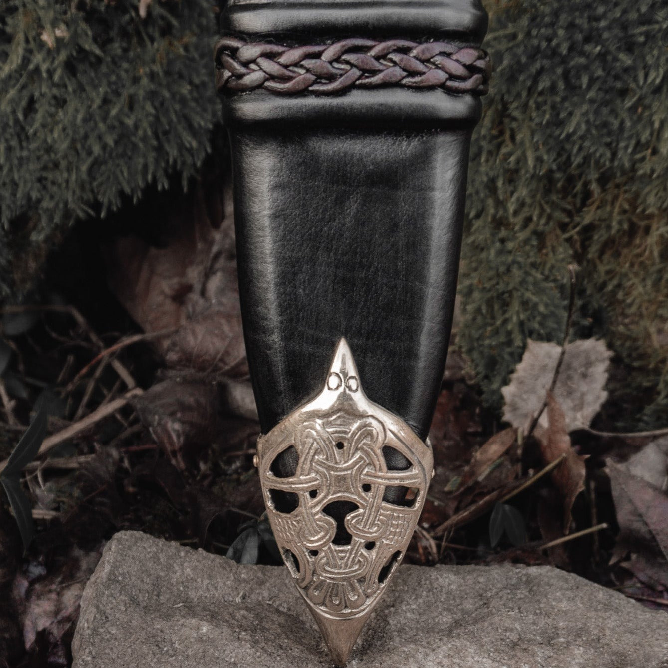 Svärdskida med flätad dekor - läderslida för svärd med vikingtida sköldsymbol vid spetsen, fotograferad mot mossiga stenar