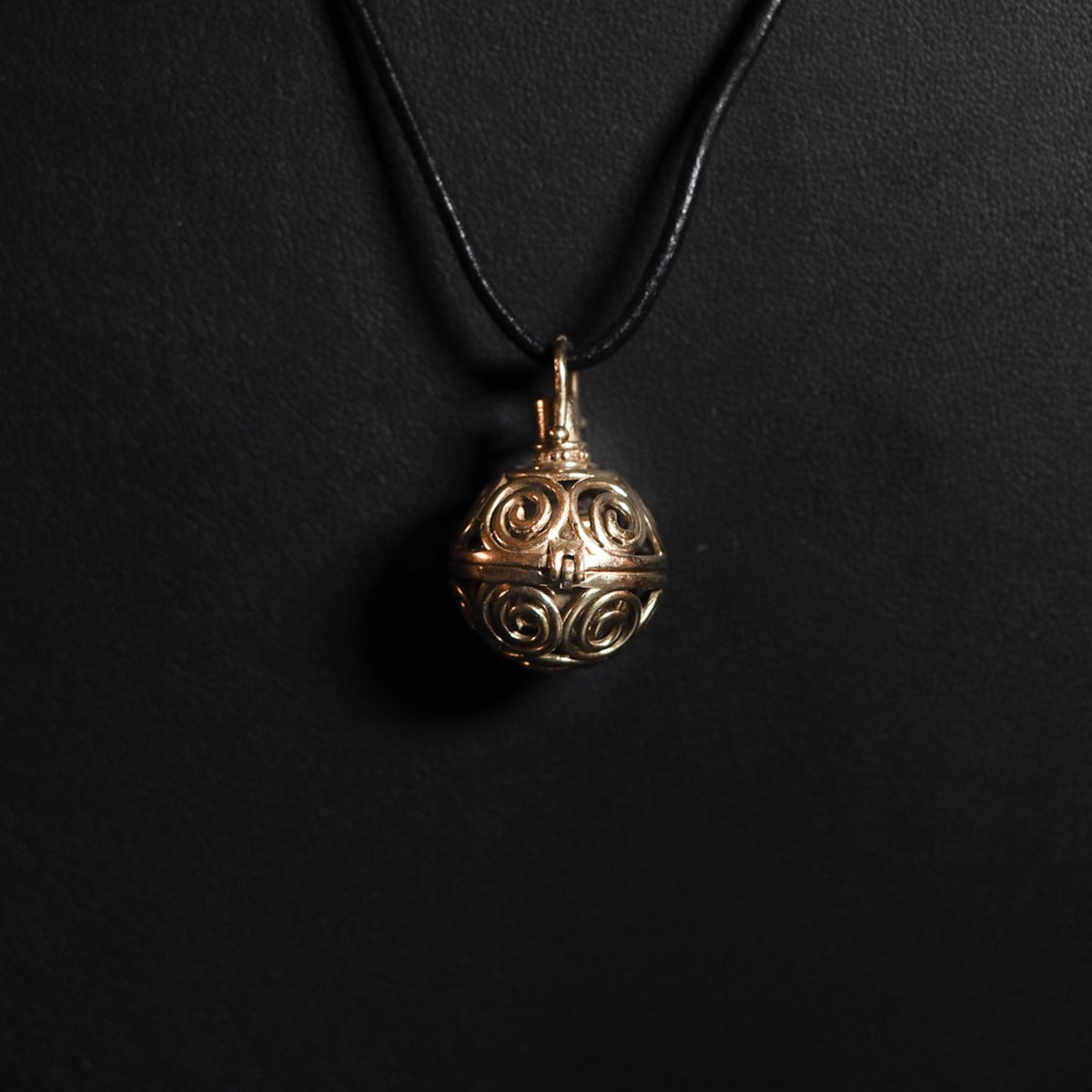 En mässing-berlock med symmetriska solspiralmönster, hängande på ett mörkt snöre. Bärlocken visar fina utskärningar av spiraler som tillsammans bildar ett solsymbol-inspirerat design. Produktfoto med närbilds-perspektiv.