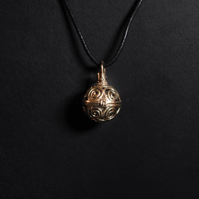 En mässing-berlock med symmetriska solspiralmönster, hängande på ett mörkt snöre. Bärlocken visar fina utskärningar av spiraler som tillsammans bildar ett solsymbol-inspirerat design. Produktfoto med närbilds-perspektiv.